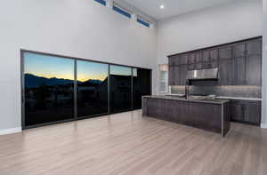 Kitchen with a towering ceiling, light wood-type flooring, backsplash, dark brown cabinets, and a center island with sink