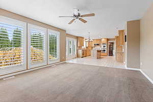 Unfurnished living room featuring light carpet, ceiling fan with notable chandelier, and a wealth of natural light