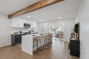 Kitchen featuring stainless steel appliances, light hardwood / wood-style flooring, beamed ceiling, white cabinets, and an island with sink