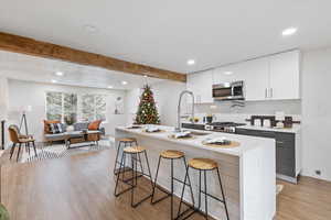 Kitchen with beam ceiling, stainless steel appliances, light hardwood / wood-style floors, and an island with sink