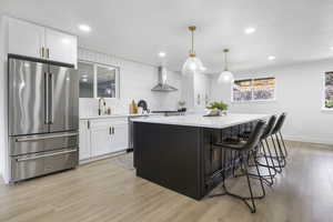 Kitchen featuring white cabinetry, a center island, hanging light fixtures, wall chimney range hood, and appliances with stainless steel finishes