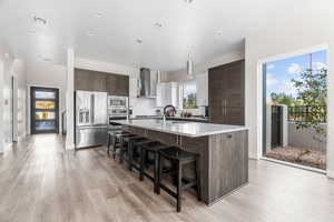 Kitchen featuring a breakfast bar, a kitchen island with sink, wall chimney exhaust hood, appliances with stainless steel finishes, and dark brown cabinets