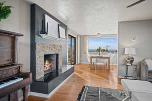 Living room with ceiling fan, a stone fireplace, light wood-type flooring, and a textured ceiling