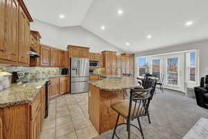 Kitchen featuring light stone counters, stainless steel appliances, vaulted ceiling, light colored carpet, and a center island with sink