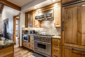 Kitchen with wall chimney exhaust hood, stainless steel appliances, tasteful backsplash, dark stone counters, and light hardwood / wood-style floors