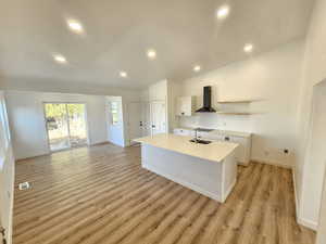 Kitchen featuring wall chimney exhaust hood, light hardwood / wood-style flooring, lofted ceiling, a center island with sink, and white cabinets