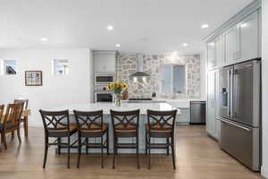 Kitchen with light wood-type flooring, gray cabinetry, stainless steel appliances, wall chimney range hood, and a center island with sink