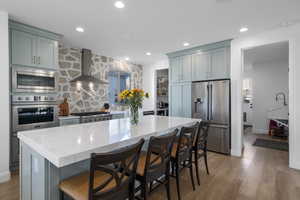 Kitchen with light stone counters, wall chimney exhaust hood, stainless steel appliances, hardwood / wood-style floors, and a breakfast bar area