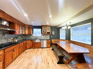 Kitchen with wall chimney range hood, sink, hanging light fixtures, stainless steel dishwasher, large windows,a breakfast nook, and a chandelier