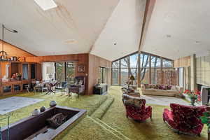 Carpeted living room with wood walls, lofted ceiling with beams, and a healthy amount of sunlight