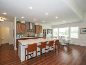 Kitchen with a kitchen breakfast bar, wall chimney range hood, an island with sink, dark hardwood / wood-style flooring, and stainless steel gas cooktop