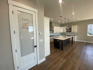 Kitchen with pendant lighting, dark wood-type flooring, a center island with sink, wall chimney range hood, and white cabinetry