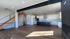 Kitchen with dark hardwood / wood-style flooring, wall chimney exhaust hood, decorative light fixtures, white cabinets, and an island with sink