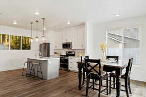 Kitchen with dark wood-type flooring, an island with sink, decorative light fixtures, white cabinets, and appliances with stainless steel finishes