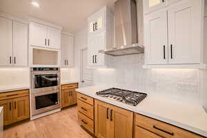 Kitchen featuring wall chimney range hood, light wood-type flooring, stainless steel appliances, and light countertops