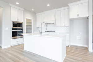 Kitchen with appliances with stainless steel finishes, premium range hood, light hardwood / wood-style flooring, white cabinetry, and an island with sink