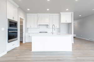 Kitchen with appliances with stainless steel finishes, tasteful backsplash, a kitchen island with sink, light hardwood / wood-style flooring, and white cabinets