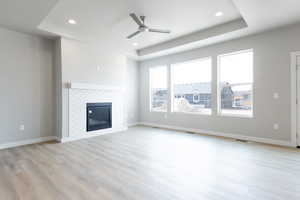 Unfurnished living room featuring ceiling fan, light wood-type flooring, a tray ceiling, and a tiled fireplace