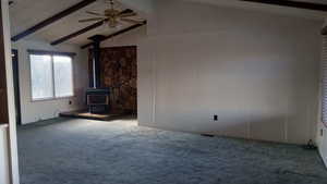 Unfurnished living room featuring lofted ceiling with beams, carpet floors, a wood stove, and ceiling fan