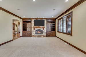 Unfurnished living room featuring built in shelves, a stone fireplace, light colored carpet, and crown molding