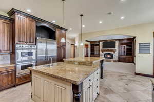 Kitchen with stainless steel appliances, light stone counters, pendant lighting, a fireplace, and a center island with sink