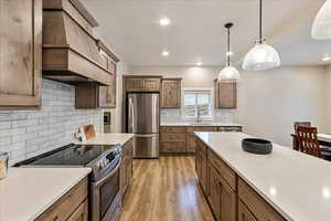 Kitchen featuring custom exhaust hood, backsplash, light hardwood / wood-style flooring, decorative light fixtures, and stainless steel appliances