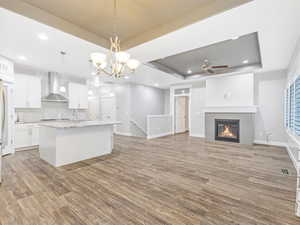 Kitchen featuring a kitchen island with sink, a raised ceiling, wall chimney range hood, hanging light fixtures, and white cabinetry