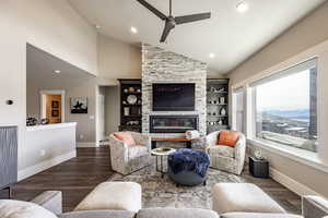 Living room with a stone fireplace, ceiling fan, high vaulted ceiling, and dark wood-type flooring