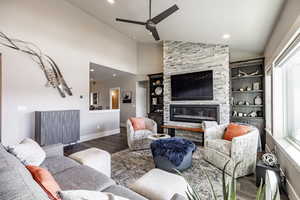 Living room featuring ceiling fan, dark wood-type flooring, built in features, high vaulted ceiling, and a stone fireplace