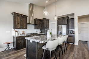 Kitchen with wall chimney exhaust hood, dark hardwood / wood-style floors, an island with sink, dark brown cabinetry, and stainless steel appliances