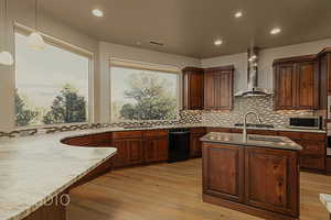 Kitchen featuring light stone counters, light hardwood flooring, pendant light fixtures, black dishwasher, and range hood