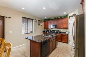 Kitchen with black appliances, sink, dark stone countertops, light tile patterned floors, and an island with sink