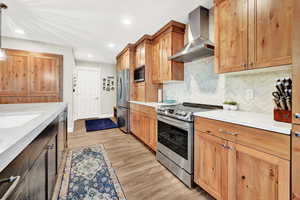 Kitchen featuring light stone counters, light hardwood / wood-style floors, wall chimney range hood, and appliances with stainless steel finishes