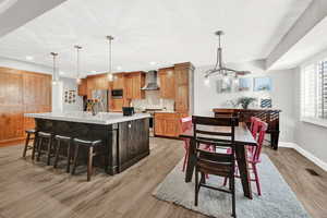 Kitchen featuring a large island with sink, wall chimney range hood, hanging light fixtures, light hardwood / wood-style flooring, and stainless steel appliances