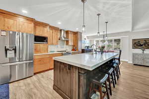 Kitchen featuring light hardwood / wood-style flooring, an island with sink, wall chimney range hood, and appliances with stainless steel finishes