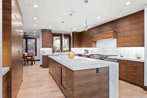 Kitchen featuring light wood-type flooring, backsplash, stainless steel gas cooktop, a kitchen island with sink, and hanging light fixtures