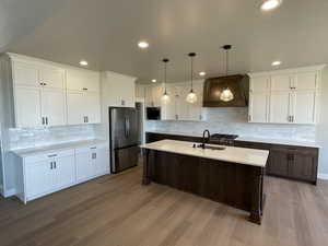 Kitchen featuring appliances with stainless steel finishes, custom exhaust hood, pendant lighting, a center island with sink, and light hardwood / wood-style flooring