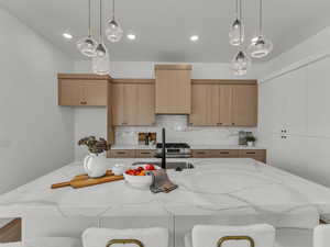 Kitchen featuring light stone counters, a sink, hanging light fixtures, decorative backsplash, and a center island with sink