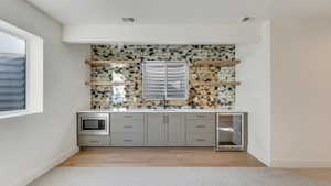 Bar featuring stainless steel microwave, backsplash, wine cooler, gray cabinets, and light wood-type flooring