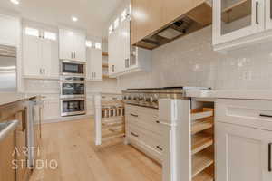 Kitchen featuring backsplash, custom exhaust hood, built in appliances, light hardwood / wood-style floors, and white cabinetry