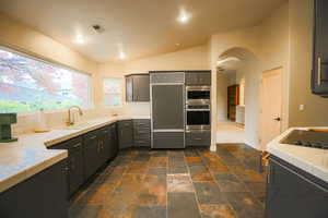 Kitchen featuring built in appliances, vaulted ceiling, tile counters, and sink