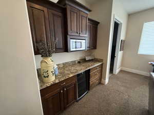 Kitchen with dark brown cabinetry, stone counters, stainless steel microwave, wine cooler, and light carpet