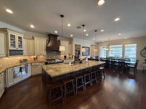 Kitchen with a large island, dark wood-type flooring, hanging light fixtures, cream cabinets, and custom exhaust hood