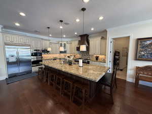 Kitchen featuring appliances with stainless steel finishes, custom exhaust hood, dark wood-type flooring, cream cabinets, and pendant lighting