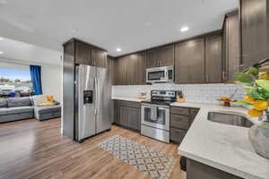 Kitchen featuring dark brown cabinetry, light stone countertops, stainless steel appliances, decorative backsplash, and light wood-type flooring