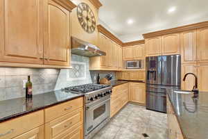 Kitchen with range hood, dark stone countertops, decorative backsplash, light tile patterned floors, and appliances with stainless steel finishes