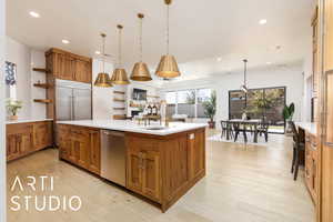 Kitchen featuring light countertops, appliances with stainless steel finishes, hanging light fixtures, brown cabinetry, and a center island with sink