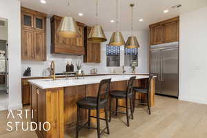 Kitchen featuring brown cabinets, a center island with sink, light countertops, glass insert cabinets, and stainless steel built in refrigerator