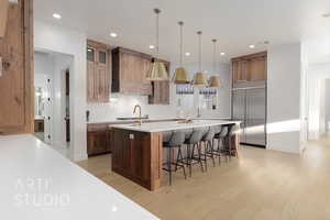 Kitchen featuring custom range hood, decorative light fixtures, stainless steel built in fridge, a center island with sink, and light hardwood / wood-style floors