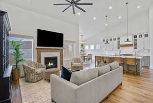 Living area featuring light wood-type flooring, ceiling fan with notable chandelier, high vaulted ceiling, and a glass covered fireplace